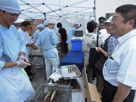 薄い水色のコックコートを着た男子生徒が網の上で魚を焼き、市長が笑顔で話しかけている写真