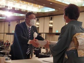 「令和3年 叙勲・褒章受章記念祝賀会」のステージ上で、グレー色の着物を着た受賞者の女性にお祝いを手渡している市長の写真