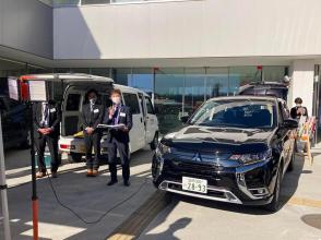 白壁の建物の前に停車した黒色の乗用車からコードが延びて、左手前に設置されたポールの上の装置と繋げて電力を供給している様子を、乗用車の傍に立つスタッフの男性が説明を行っている写真