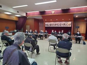 前方に「市長と語ろうタウンミーティング」と書かれた横断幕が掲げられた会場で、2列に設置された長机の席に座った関係者と間隔を開けて設置されたパイプ椅子に座った参加者が向かい合って座っている新里会場の様子を後方から撮影した写真