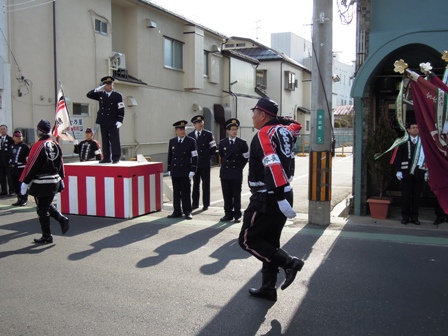 紅白幕が付けられた小さなステージに敬礼している男性が立ち、その前の道路を宮古と書かれた黒い半被をきた消防団員の男性達が歩いている写真