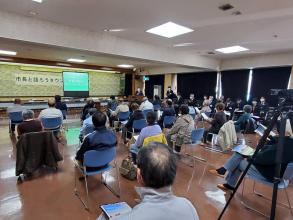 小さなステージの幕に「市長と語ろうタウンミーティング」と書かれた横断幕とスクリーンが設置された田老会場で、右の窓側に関係者、間隔を開けて設置されたパイプ椅子に参加者が座り、右前方に設置された檀上に立つ市長の話を参加者が聞いている様子を後方から撮影した写真
