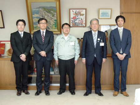 作業着を着た市長とスーツを着た男性4名が横並びで記念撮影している写真