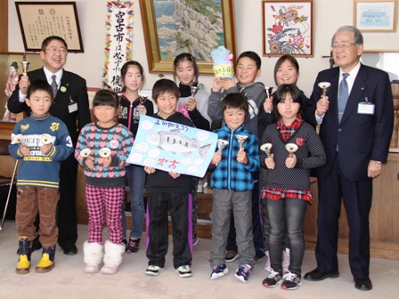 魚が描かれた絵を手に持つ男の子と、ハンドベルを持つ市長と小学生の子供たちが横並びに並んでいる写真