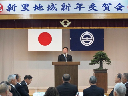 「新里地域新年交賀会」の横断幕の元、演壇に立つ市長と、椅子に座って話を聞く参加者の写真