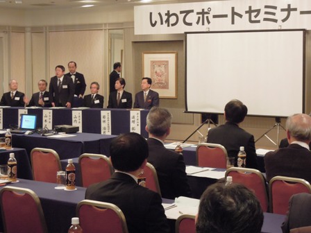 「いわてポートセミナー」と書かれた吊り看板があり、舞台上の来賓席に市長と関係者がいる様子を、参加者側から見た写真