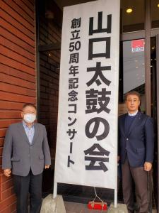 入り口に飾られた山口太鼓の会 創立50周年記念コンサートの看板をはさんで市長と佐々木清会長が並んで記念撮影をしている写真