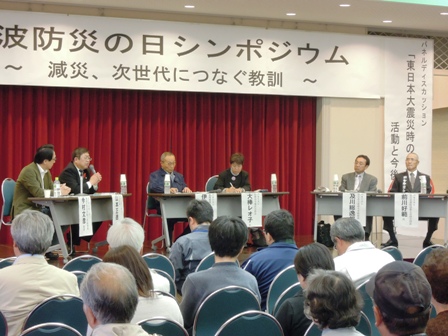 「津波防災の日シンポジウム ～減災、次世代につなぐ教訓～」の吊り看板の下、市長と関係者が舞台上でパネルディスカッションをしており、観客側が聴衆している写真