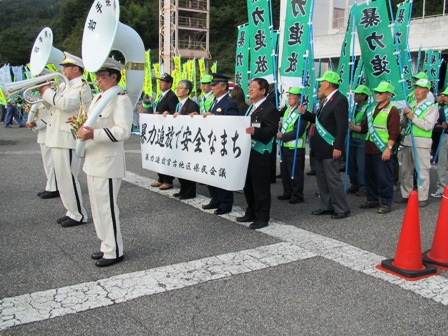白い制服を着てスーザフォンなどの管楽器を持つ3名の音楽隊の方の後ろで、横断幕やのぼり旗を持ち整列する市長と関係者の方々の写真