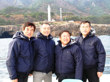 同じジャンパーを着用した市長を含む4名が遠くに見える灯台を背景に海上で記念撮影をしている写真