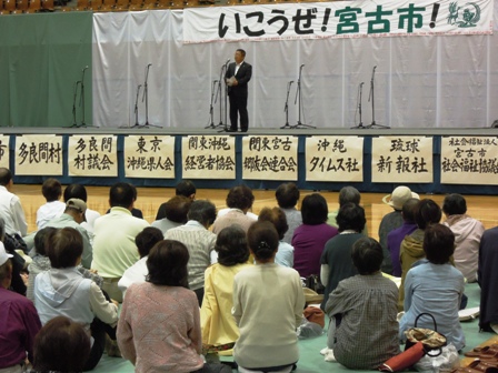 「いこうぜ！宮古市！」と書かれた横断幕の前にマイクスタンドが5つ並べられ、真ん中のマイクスタンドでスーツをきた男性が話しているのを参加者の方達が聞いている写真