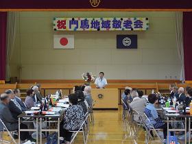 ステージに「祝門馬地域敬老会」の吊り看板の下、長机が並べられた席に参加者が座っており、演壇で挨拶をする市長の写真