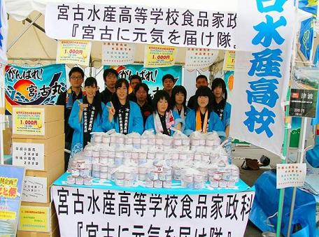 青色の半被を着た宮古水産高校の生徒がテントの下で机の上につまれている特製缶詰を販売している写真