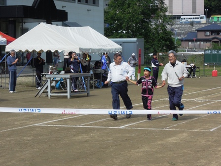 小学校のグランドで市長と小学生くらいの子供と参加者が3人で手をつないで走っており、まもなくゴールテープを切ろうとしている写真