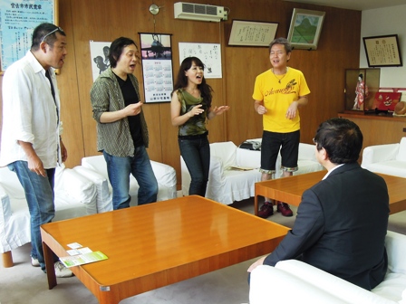 白い布におおわれた椅子に市長が座り、男女4名が市長と向い合わせに立って歌を歌っている様子の写真