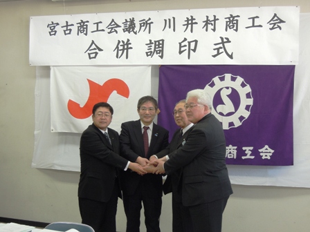 「宮古商工会議所川井村商工会合併調印式」と書かれた横断幕の下で男性4名が両手で握手をし記念撮影している写真