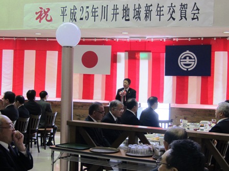 壁に紅白幕が張られており「平成25年川井地域新年交賀会」の横断幕の下、マイクスタンドの前に立つ市長と、椅子に座り話を聞く参加者の写真