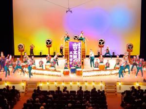 舞台上で2名の人が座って演奏している大きな太鼓が中央に設置され、その後ろに5個の太鼓、前にもいろいろな種類の太鼓が設置されて演奏している様子を写した写真