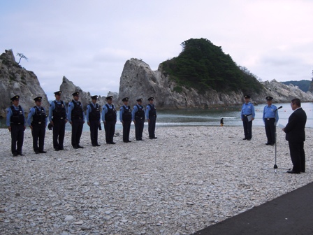 海辺の砂利の上に9名の警官が並び、向かい合わせでスーツを着た男性がマイクスタンドを使い話をしている写真
