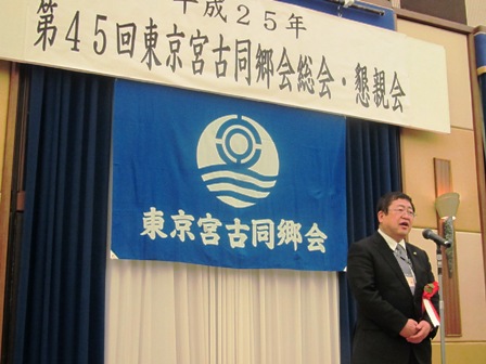 「第45回東京宮古同郷会総会・懇談会」の吊り看板と「東京宮古同郷会」と書かれた同郷旗が壁面にある舞台で、スタンドの前に立つ市長の写真