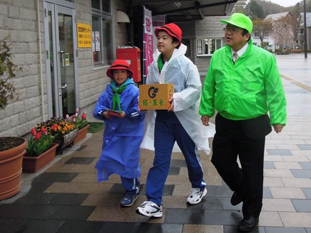 黄緑色のウインドブレーカーを着た市長と、募金箱を持ったレインコート姿の子供たちが宮古駅を歩いている写真