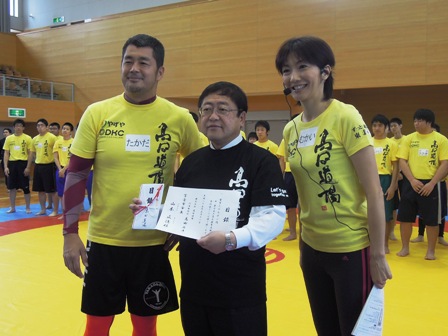 高田道場と書かれた黄色のTシャツを着た参加者達の前で、目録を手に持つ市長と、高田延彦さんと向井亜紀さんが並んでいる写真