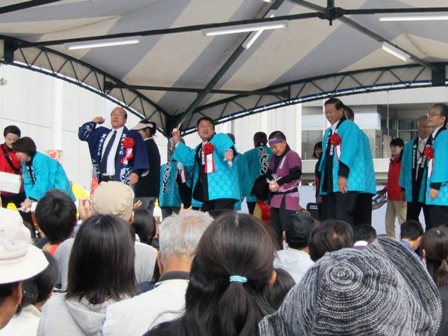 青い法被を着た市長と関係者の方々が、舞台下にいる参加者たちに向け餅まきをしている様子の写真