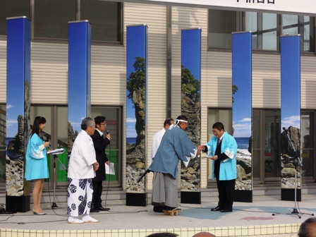 エントランスのような場所で、水色や白の羽織を着た参加者が市長と握手をしている写真