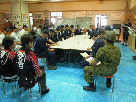 ブルーシートが敷かれ机と椅子が置かれた場所に迷彩服をきた自衛隊の方や警察官、宮古と書かれた半被をきた方々集まり話し合いをしている写真