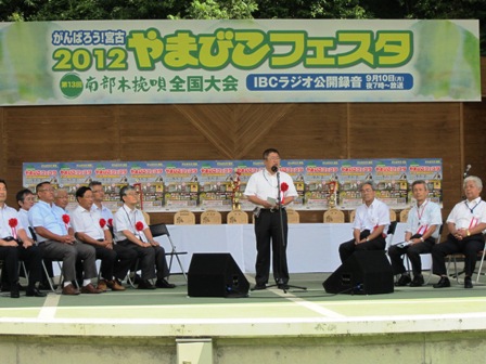 「がんばろう！宮古2012やまびこフェスタ南部木挽唄全国大会」書かれた横断幕が掲げられたステージの上で胸に花飾りをつけたスーツ姿の男性達がパイプ椅子に座り、中央に置かれたマイクスタンドの前に立つ男性が話をしている写真