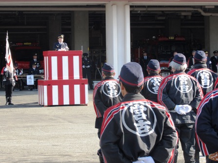 宮古消防署で紅白幕の演壇に立つ市長と、消防はっぴを着た消防団員が整列している出初式の写真
