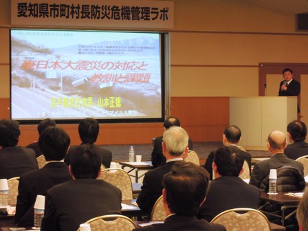 「愛知県市町村長防災危機管理ラボ」の横断幕の下、「東日本大震災の対応と教訓と課題」と題した資料がスクリーンに映し出されており、演壇で講演をする市長と、話に耳を傾ける参加者の写真