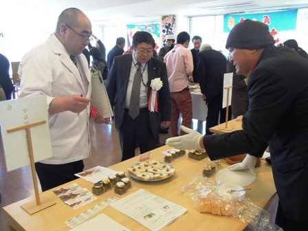 机の上に資料と加工食品が皿の上に並べられており、商品の説明を受ける様子の市長と関係者の写真
