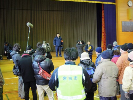 防寒服を着た参加者や関係者が、体育館の舞台に立つ市長を見ている写真