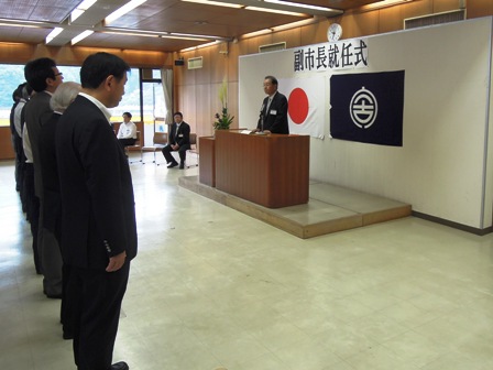 「副市長就任式」と書かれたバックパネルの前で演壇に立つ山口公正副市長と向い合せに、横一列に並び話を聞く関係者の写真