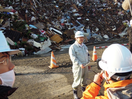 震災で発生したがれきが積まれた山の前に、防災服にヘルメットをかぶった市長が、作業者に向けて話をしている様子の写真