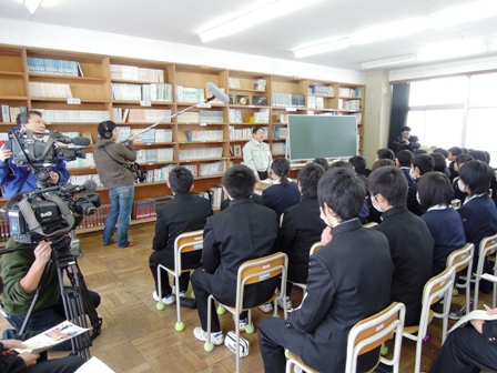 天井まである本棚の前で作業着を着た市長が話しているのを椅子に座って聞いている生徒達の写真