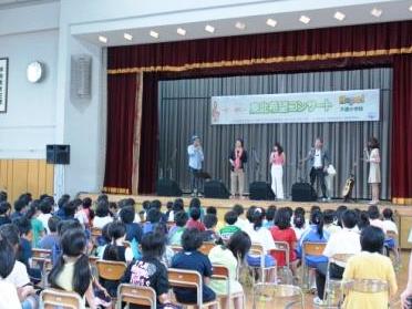 体育館のステージ上に「東北希望コンサート」と書かれた横断幕があり、男女5名が歌を歌っているのをステージ下で子供達が椅子に座り聞いている写真