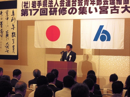「岩手県法人会連合会青年部会連絡協議第17回研修の集い宮古大会」と書かれた横断幕の下に日の丸と宮古市市章の旗が飾られ、その前の演壇で話しているのを参加者たちが座って聞いている写真