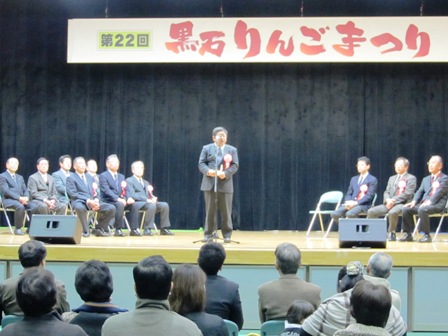 「黒石りんごまつり」の吊り看板があり、ステージ両側に関係者が椅子に座る中央にマイクスタンドの前に立つ市長と、その話を聞く参加者の写真