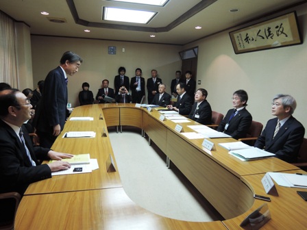 円形の茶色いテーブルの周りに着席する市長と関係者の男性たち、起立する男性1名を写した写真