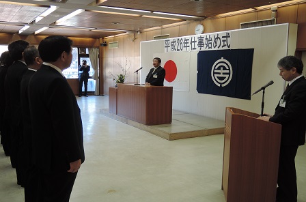 「平成26年仕事始め式」と書かれた紙が貼られた壁の前に立ち、演台に置かれたマイクに向かい話をする市長の写真