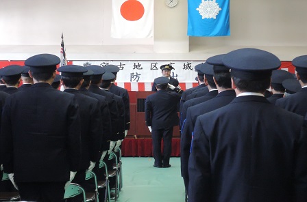 パイプ椅子の前に立ち整列する制服を着た消防職員の方々の前で、右手で敬礼をする男性と、向かい側に立つ市長の写真