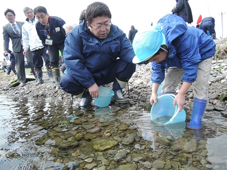 青いバケツを川につけて稚魚の放流を行う様子の男の子と、その横でしゃがみこみ様子を見ている市長の写真