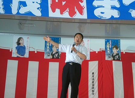 紅白幕とポスターが貼られている壁の前で、右手を大きく横に広げて左手に持ったマイクで話をする市長の写真