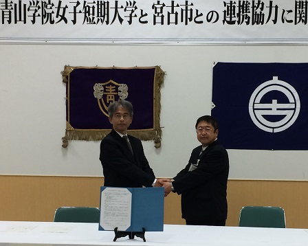 校旗や宮古市の市町村旗が掲げられた壁の前で両手で握手をする市長と男性の写真