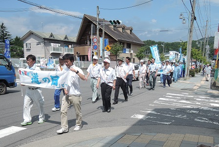 「道の日」と書かれた横断幕を持つ2名の男性を先頭に、整列して道路を更新する様子の市長と関係者の方々の写真