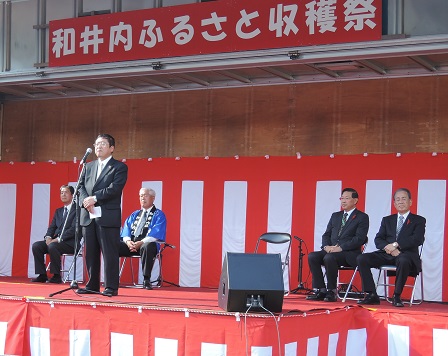 「和井内ふるさと収穫祭」と書かれた横断幕と紅白幕が掛けられた舞台上で、スタンドマイクに向かい話をする市長の写真