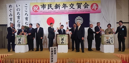 市民新年交賀会と書かれた横断幕があるステージで、3個の酒樽をそれぞれ4人で 鏡開きをしている写真