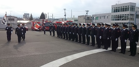 制服姿で整列している消防署職員の方々の前を、旗を持った職員も同行した制服姿の市長が歩いている写真
